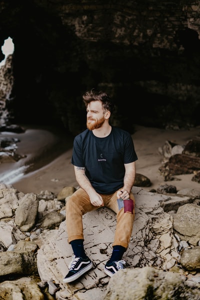 Dressed in a black round collar T-shirt man sat on a rock and brown trousers
